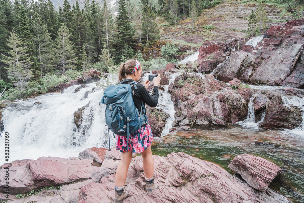 Woman hiker with a big backpack takes photos of Redrock Falls waterfall in Glacier National Park