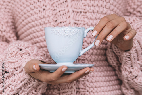 A girl with a fashionable pink manicure holds a blue mug against the background of a knitted pink sweater