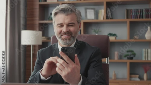 Smiling middle-aged bearded businessman using smartphone while sitting in office. A stylish businessman is using a mobile phone at his workplace.