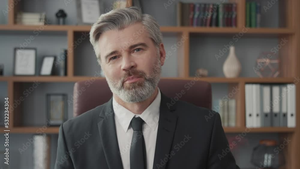 Successful middle-aged businessman greeting someone via online video call on his laptop while sitting at his desk and working in his office. Medium shot of smiling confident man talking to camera