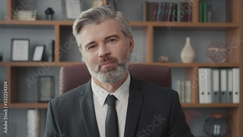 Successful middle-aged businessman greeting someone via online video call on his laptop while sitting at his desk and working in his office. Medium shot of smiling confident man talking to camera