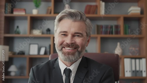Close-up portrait of a successful gray-haired businessman in a suit sitting at a desk with a laptop in the office. Portrait of a confident middle-aged man. Smiling and looking at the camera