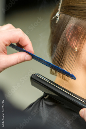 Hairstylist is straightening short hair of young brunette woman with a flat iron in a hairdresser salon, close up