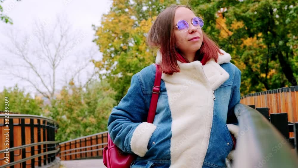 Young beautiful woman in glasses with short hair in warm denim jacket looking away, standing on footbridge in cool weather. Young beautiful woman in glasses with short hair in warm denim jacket looking away, standing on footbridge in cool weather.