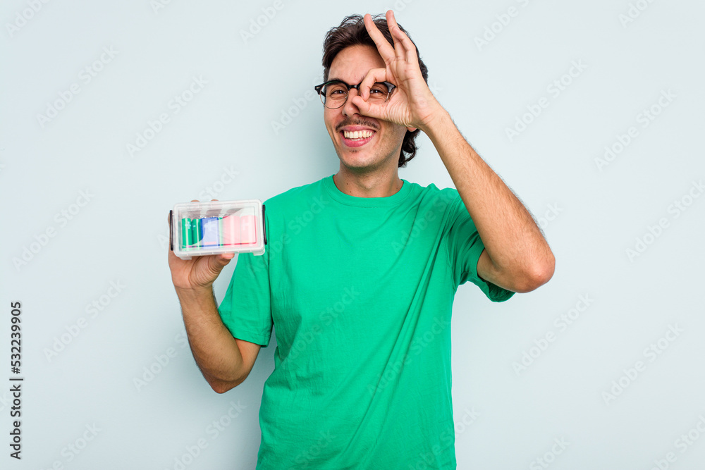 Young hispanic man holding battery box isolated on white background excited keeping ok gesture on eye. Young hispanic man holding battery box isolated on white background excited keeping ok gesture on eye.