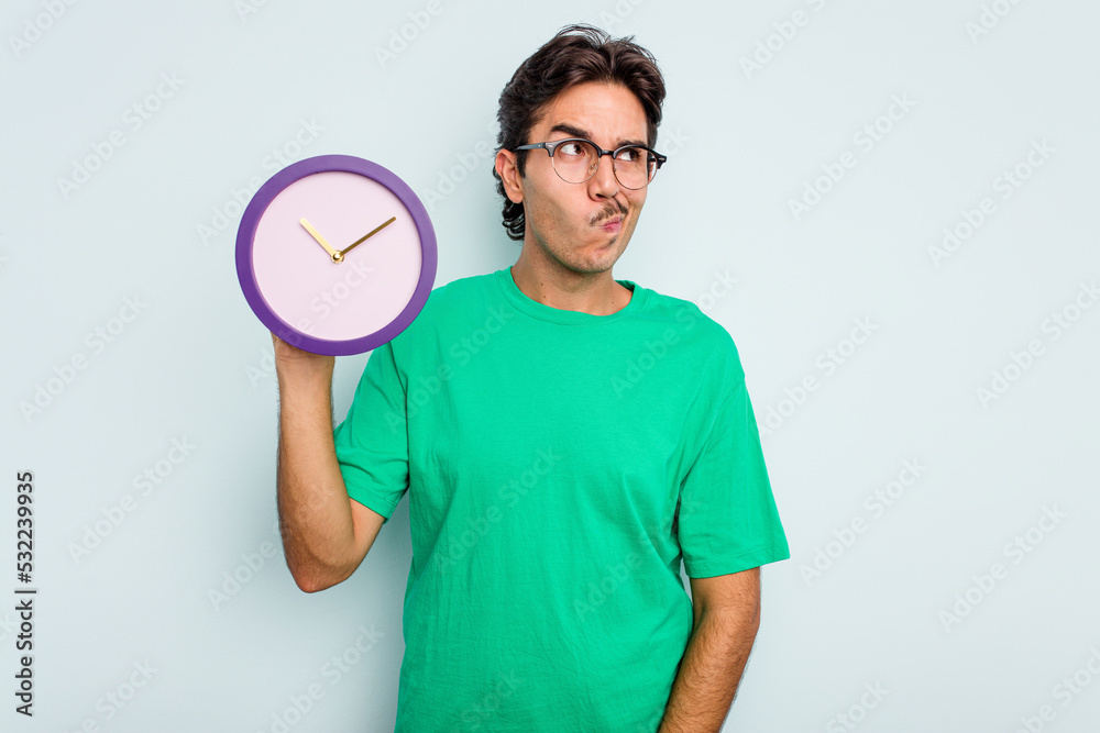 Young hispanic man holding a clock isolated on white background confused, feels doubtful and unsure. Young hispanic man holding a clock isolated on white background confused, feels doubtful and unsure.