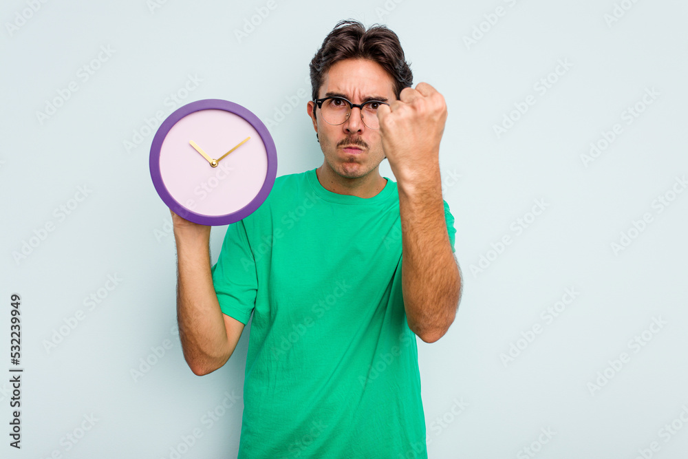 Young hispanic man holding a clock isolated on white background showing fist to camera, aggressive facial expression. Young hispanic man holding a clock isolated on white background showing fist to camera, aggressive facial expression.