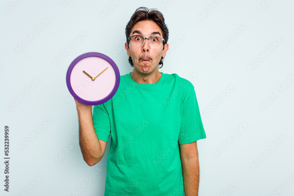 Young hispanic man holding a clock isolated on white background shrugs shoulders and open eyes confused. Young hispanic man holding a clock isolated on white background shrugs shoulders and open eyes confused.