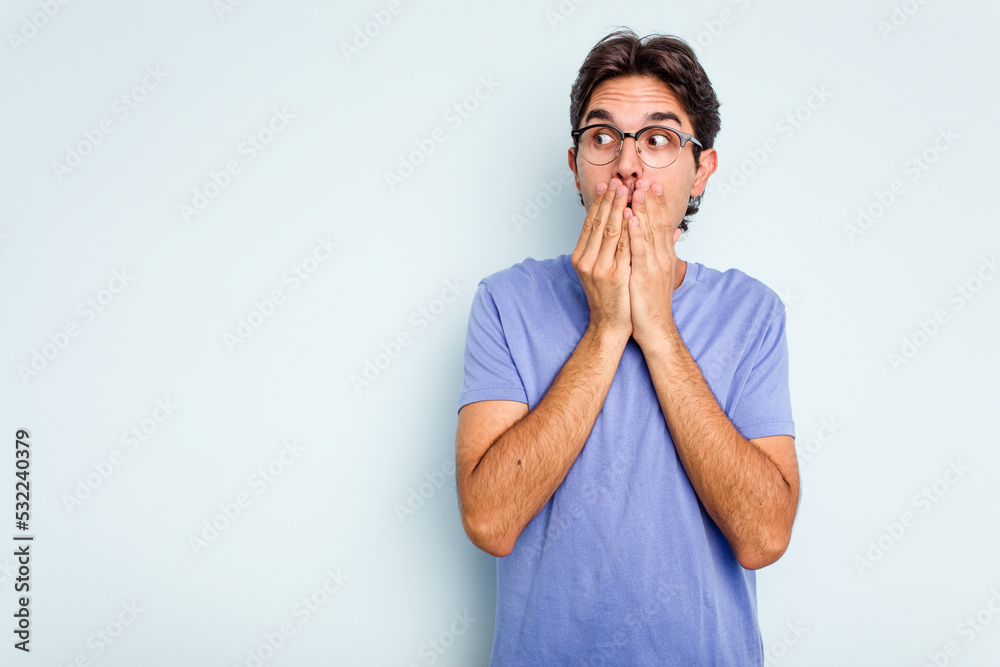 Young hispanic man isolated on blue background thoughtful looking to a copy space covering mouth with hand.