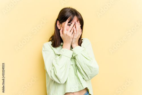 Young caucasian woman isolated on yellow background blink at the camera through fingers, embarrassed covering face.