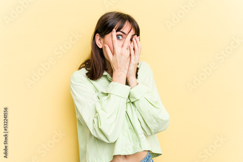 Young caucasian woman isolated on yellow background blink through fingers frightened and nervous.