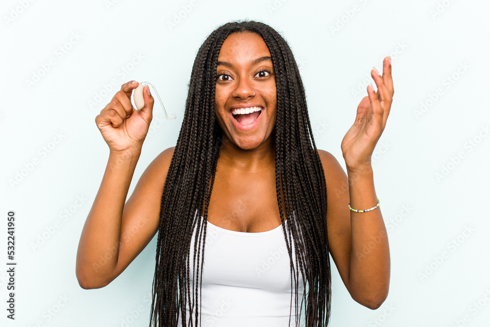 Young African American woman holding hearing aid isolated on blue background receiving a pleasant surprise, excited and raising hands. Young African American woman holding hearing aid isolated on blue background receiving a pleasant surprise, excited and raising hands.