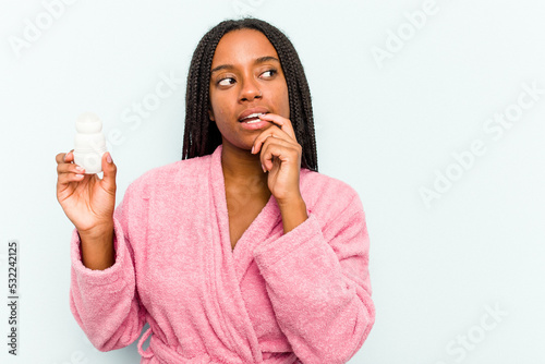 Young African American woman wearing bathrobe holding a deodorant isolated on blue background relaxed thinking about something looking at a copy space.