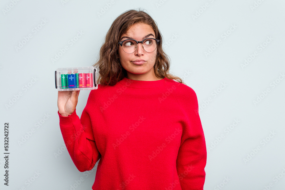 Young caucasian woman holding battery box isolated on blue background confused, feels doubtful and unsure. Young caucasian woman holding battery box isolated on blue background confused, feels doubtful and unsure.