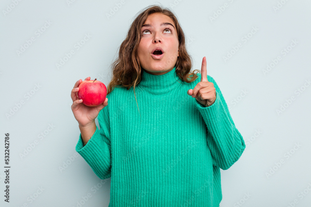 Young caucasian woman with an apple isolated on blue background pointing upside with opened mouth. Young caucasian woman with an apple isolated on blue background pointing upside with opened mouth.