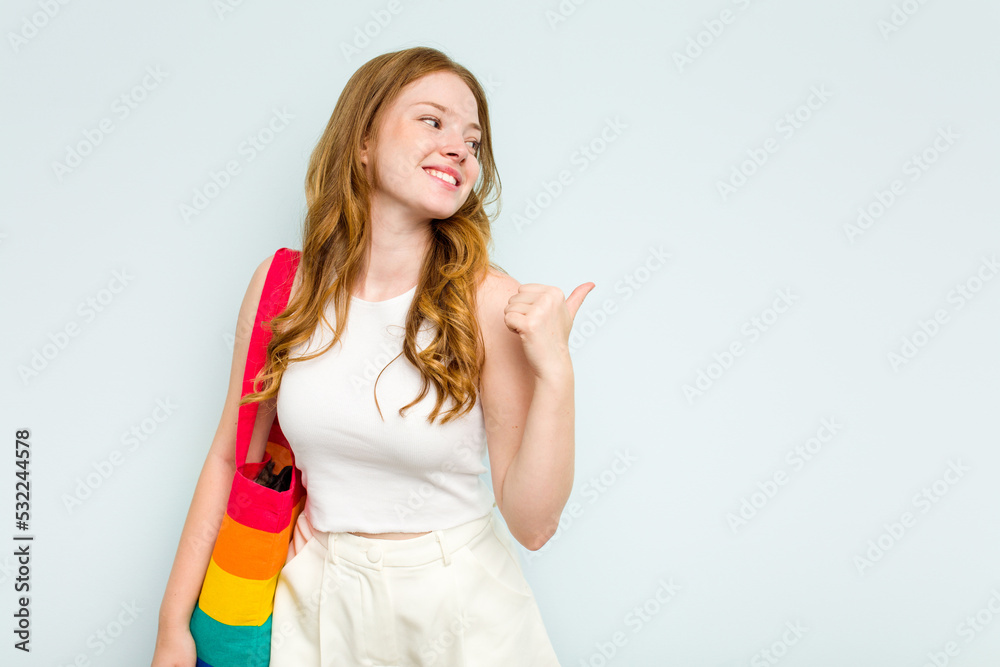 Young caucasian woman holding LGTBI bag isolated on blue background points with thumb finger away, laughing and carefree. Young caucasian woman holding LGTBI bag isolated on blue background points with thumb finger away, laughing and carefree.