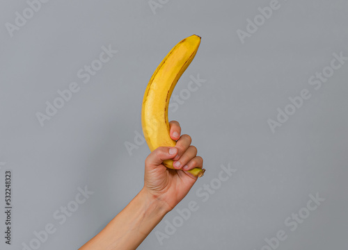 A woman's hand is holding a yellow ripe banana. Weapon symbol. Gray background.