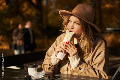 a beautiful young woman sits in an outdoor cafe at a wooden table in an autumn park, and eats kebab or shawarma
