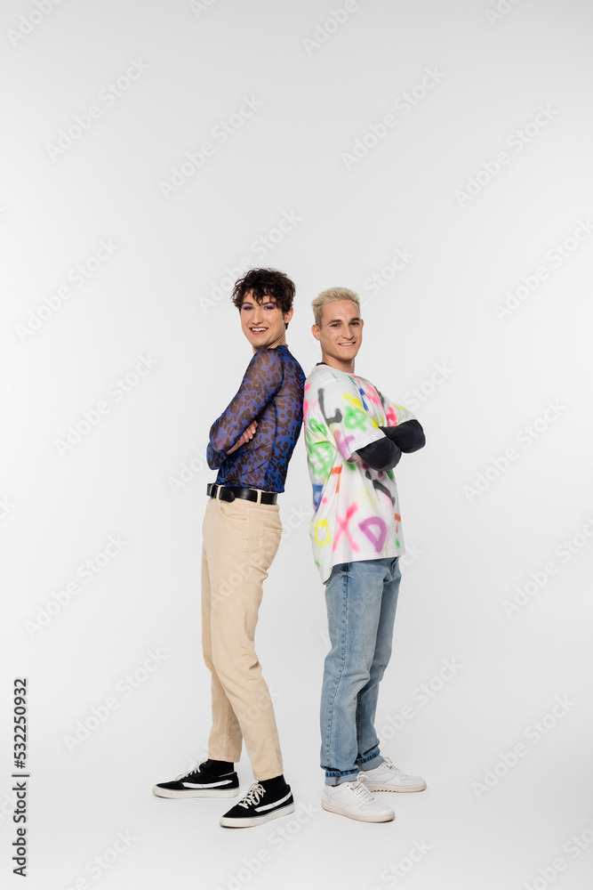 full length of cheerful gay man and queer person standing back to back with crossed arms on grey background. full length of cheerful gay man and queer person standing back to back with crossed arms on grey background.