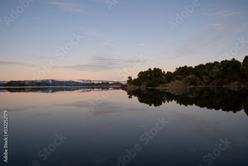 Panorama view of the peaceful lake, forest, cliffs and mountains at sunset. Beautiful orange and blue colors in the sky and water surface.