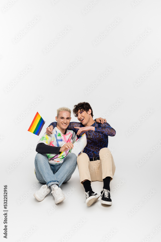 Fototapeta premium smiling gay man with small lgbtq flags sitting near queer person pointing with finger on grey background.