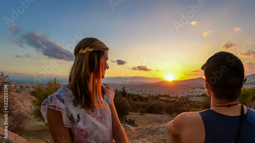 Young couple watching sunset over city of Athens seen from Filopappou Hill (hill of muses), Athens, Attica, Greece, Europe. Athens cityscape and Aegean sea. Beautiful sunset point with aerial view