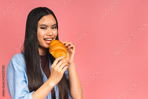 Portrait of young beautiful hungry asian woman eating croissant. Isolated portrait of woman with fast food over pink background. Diet breakfast concept.