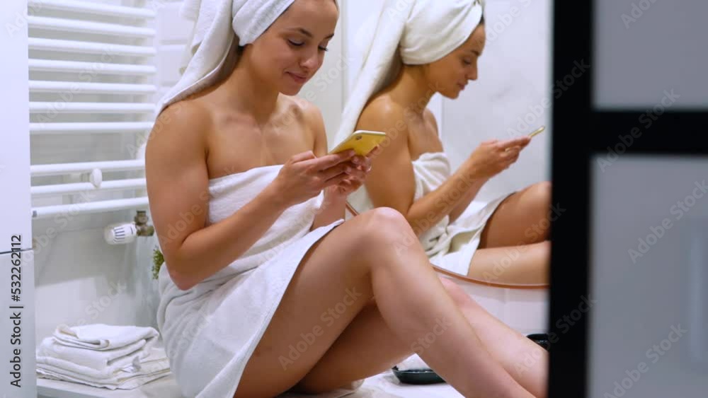 Caucasian young beautiful woman in towel sitting in bathroom texting on smartphone after taking shower in good mood. Someone peeping on lady in bathroom at home using cellphone