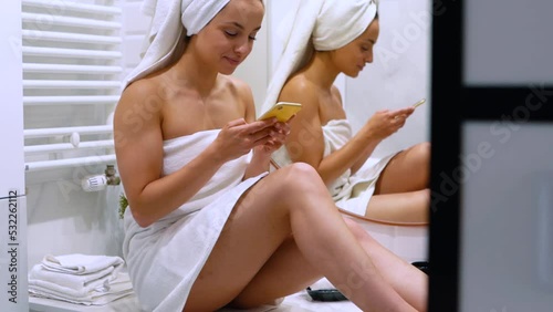 Caucasian young beautiful woman in towel sitting in bathroom texting on smartphone after taking shower in good mood. Someone peeping on lady in bathroom at home using cellphone
