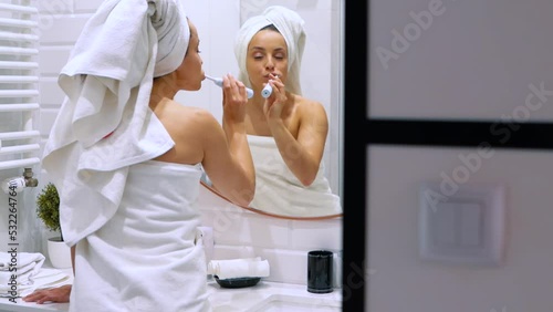 Rear of young happy woman standing in bathroom after shower brushing her teeth with an electric toothbrush. Beautiful Caucasian attractive lady having morning routing at home
