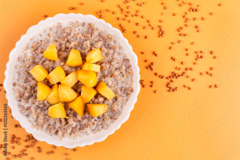 Buckwheat porridge with milk and juicy peach in a white bowl (plate) on an orange background. Porridge for a child (baby). Healthy breakfast. Space for text. Buckwheat porridge with milk and juicy peach in a white bowl (plate) on an orange background. Porridge for a child (baby). Healthy breakfast. Space for text.