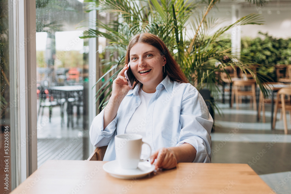 Smiling woman talking on the phone and drinking coffee or tea indoors on a rainy day. Young cheerful lady having fun during a funny conversation call. Concept of easy breakfast. Smiling woman talking on the phone and drinking coffee or tea indoors on a rainy day. Young cheerful lady having fun during a funny conversation call. Concept of easy breakfast.