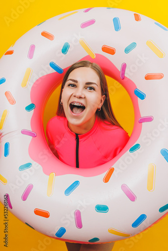 Beautiful young woman in a stylish pink swimsuit with a donut inflatable ring on a yellow background