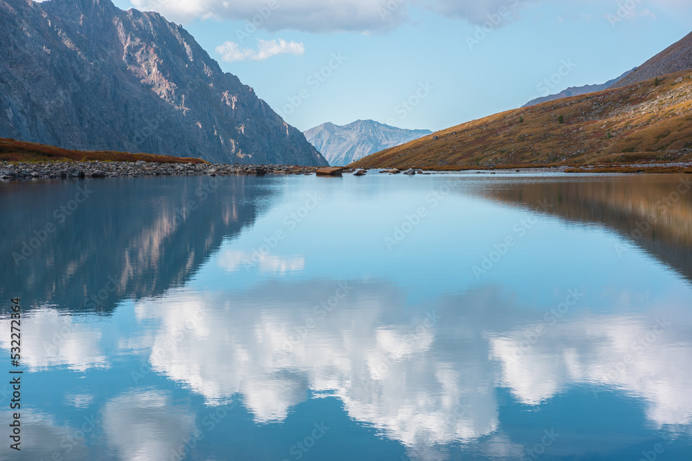 Tranquil autumn landscape with clouds reflection on smooth mirror surface of mountain lake in high hanging valley. Meditative view from calm alpine lake to mountain vastness. Glacial lake clear water.