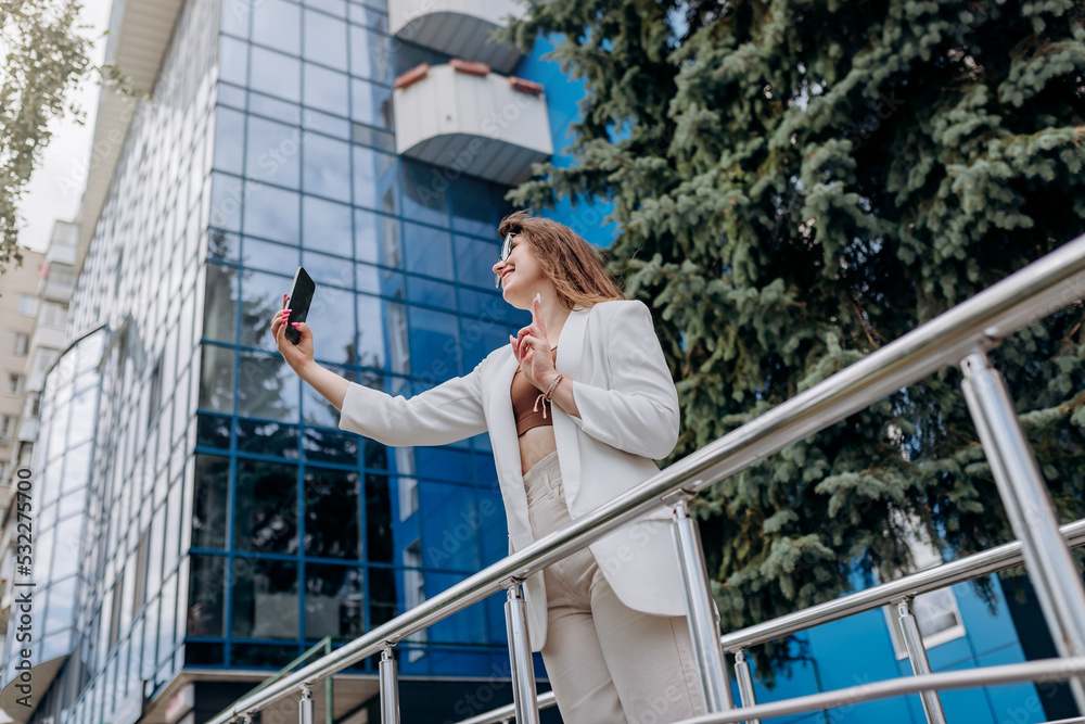 Smiling business woman in white suit and sunglasses making video call to friend using phone during break standing near modern office building