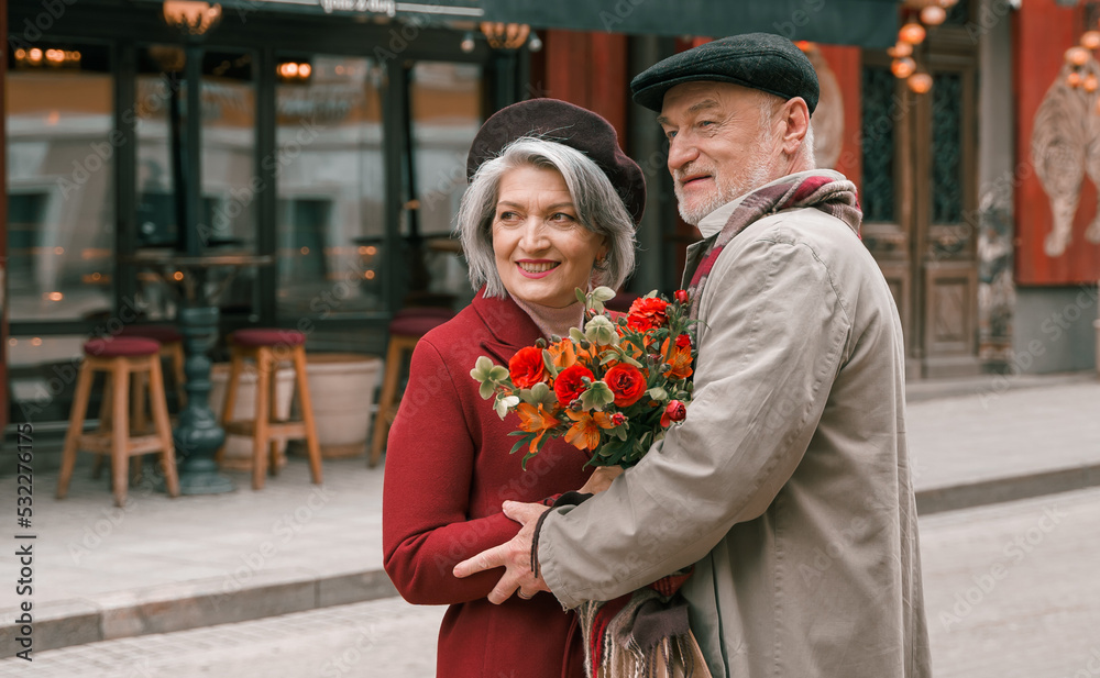 Obraz premium Elderly senior love couple. Old retired man woman together on romantic date.Aged husband wife walking on city street with flowers.Stylish elder hugging people pensioner in red coat.Happy family years