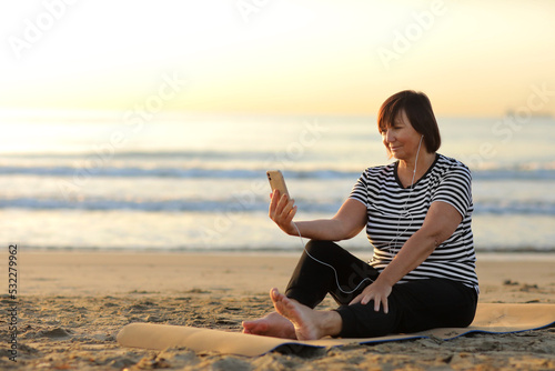 middle ages woman takes a break while sitting and relaxing on the yoga fitness mat and talking on smartphone with headphones on the beach by the sea. outdoor workout. Healthy, chill out lifestyle.