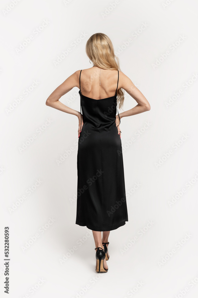 Naklejka premium Elegant beautiful slender blonde girl in a fashion long black dress with heels shoes stands back to the camera in the studio. Party female style, fashion and beauty
