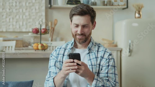 Happy handsome Caucasian man chatting on social networks, watching social media videos and having fun with smartphone in cozy living room at home. A middle-aged man is resting on a comfortable sofa.