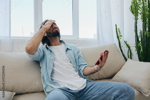 A young man looks angrily and sadly at the phone screen at home sitting on the couch. Bad news and bad luck at work