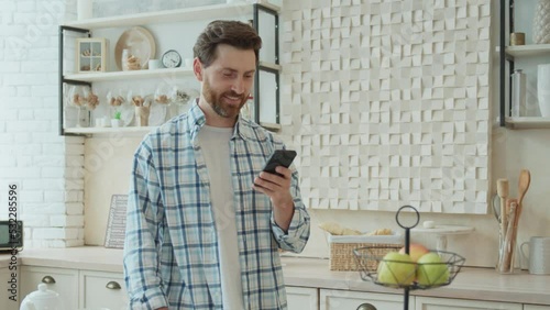 Portrait of a handsome bearded middle-aged man texting on the phone in his kitchen. Close-up of a smiling man's face. Pleasant communication with friends online, creating a post in a social network