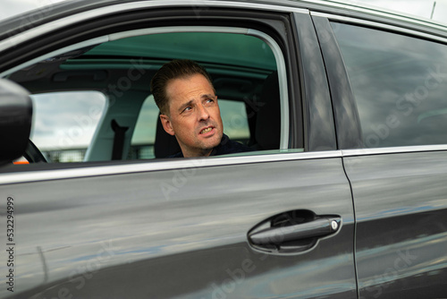 White Caucasian curious man is sitting on the drivers side of a gray black modern car and is looking outside through an open car window. There are no trademarks in the shot. © Jeffrey