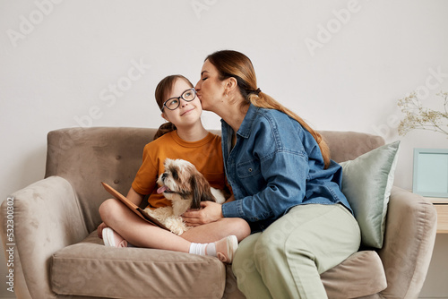 Minimal portrait of caring mother kissing daughter with Down syndrome on cheek while sitting together on couch at home © Seventyfour
