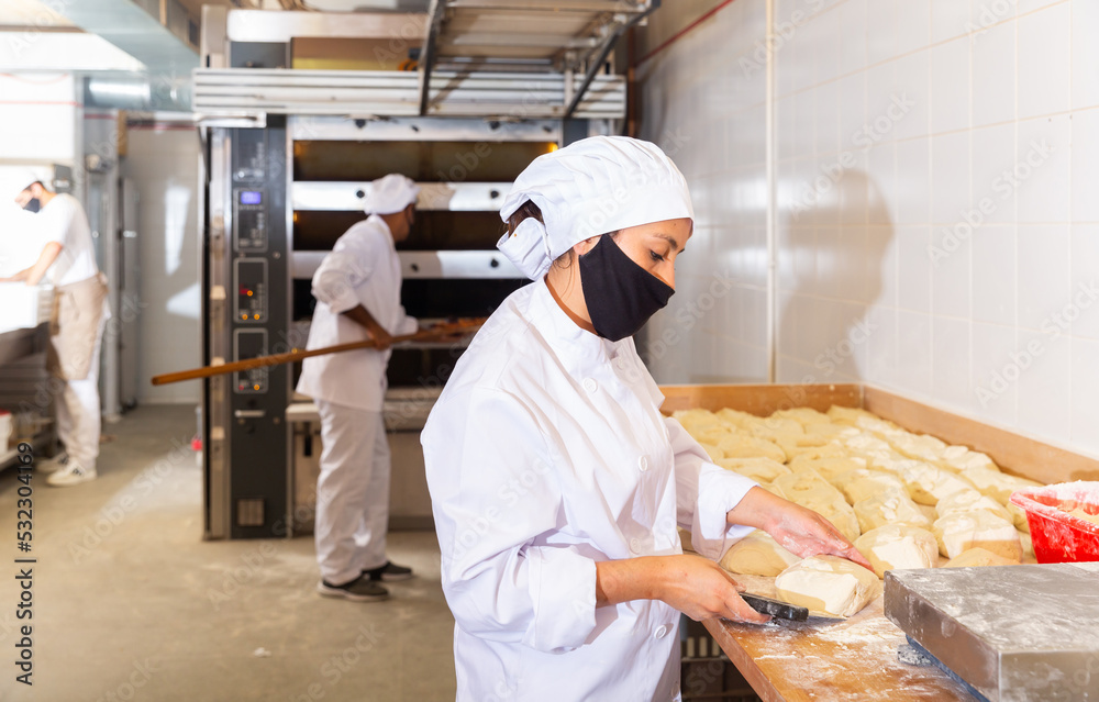 Female bakery worker wearing protective mask cuts dough with knife into identical loaves in bakery kitchen