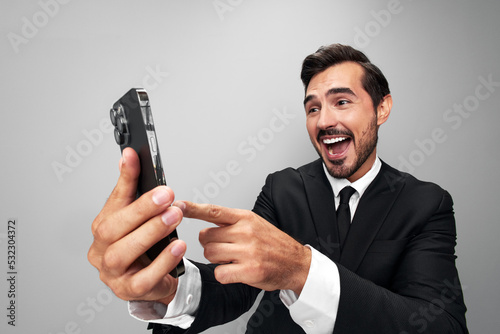 Man businessman in suit takes selfies on phone posing in front of smartphone camera with smile with teeth happy win on gray background close-up face wide camera angle