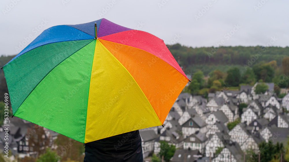 Woman standing on a hill with a rainbow umbrella on a rainy day and looking at a old town with half-timbered houses