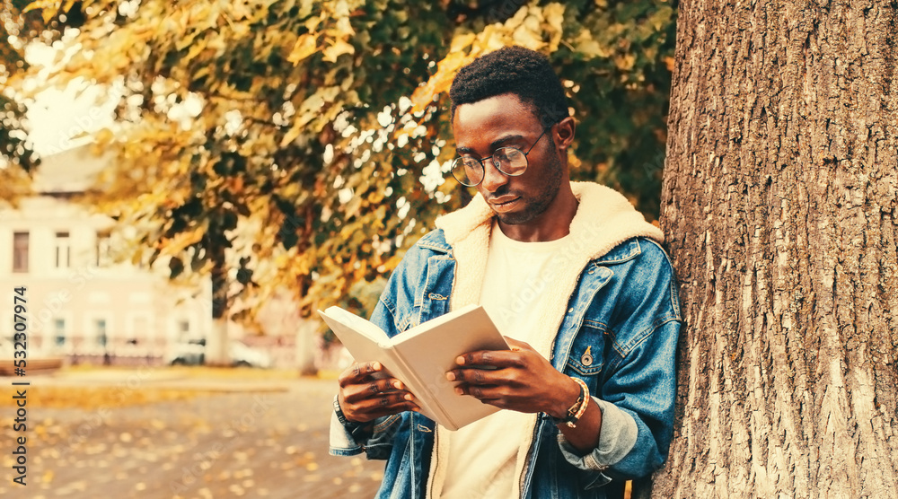 Fototapeta premium Portrait of young african man student reading a book wearing eyeglasses in autumn city park