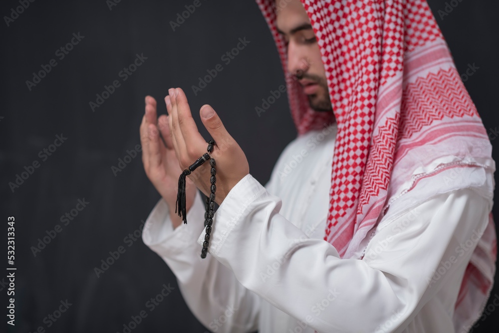 A young Arabian man in traditional clothes making a traditional prayer to God keeps his hands in praying gesture in front of a black background
