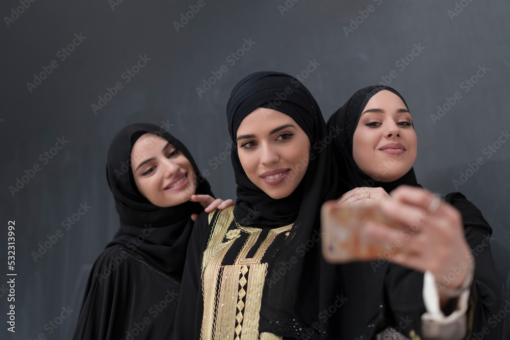 Group of young beautiful muslim women in fashionable dress with hijab using smartphone while taking selfie picture in front of black background