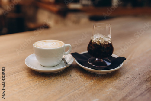 Fototapeta Naklejka Na Ścianę i Meble -  Ice coffee in a tall glass with whiskey and latte coffee in a white cup with a plate on  a wooden table. Cold and hot summer drink on a dark napkin and white plate, light wooden table in café